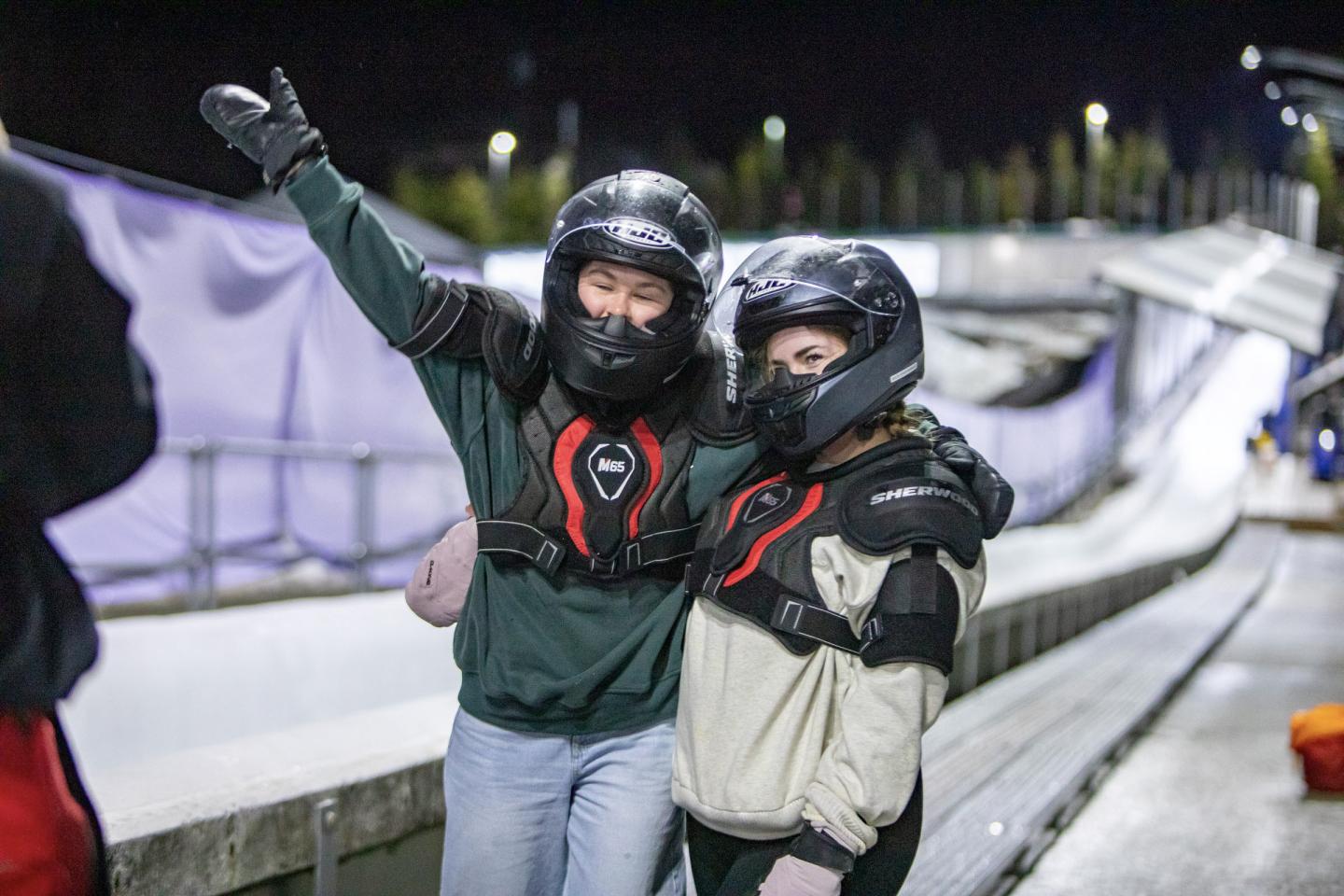 Two people in helmets pose next to a bobsleigh track at night, one arm raised in excitement.