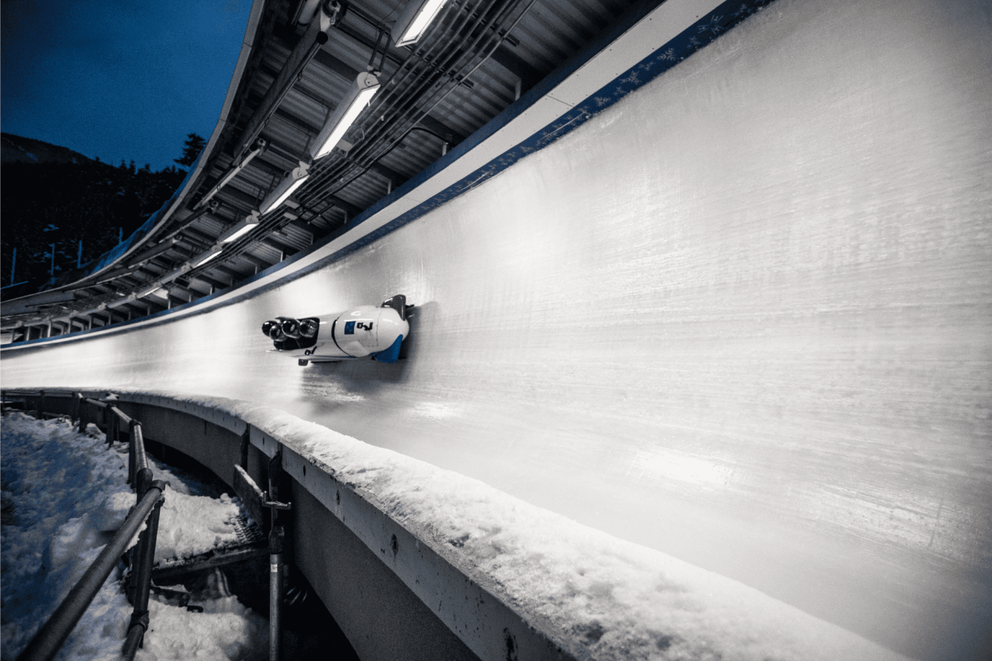 Bobsled racing down an icy track at night, under bright lights.