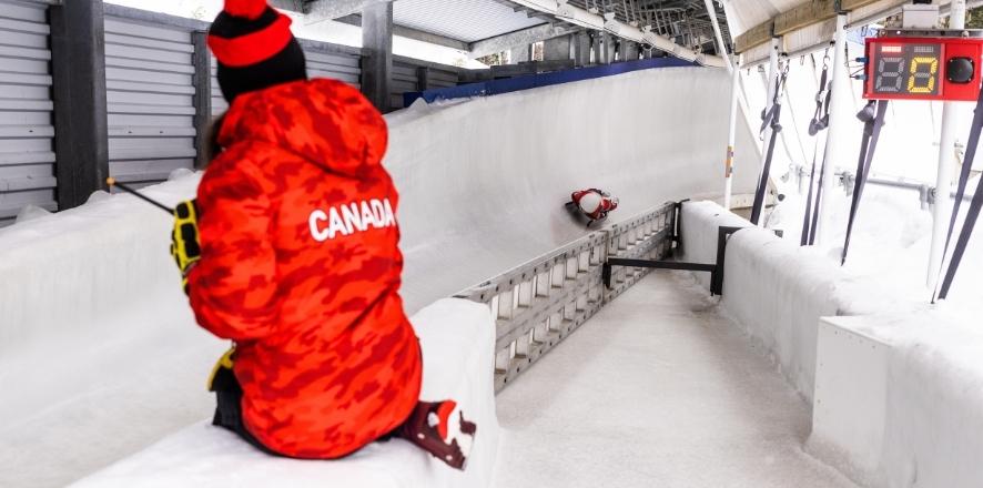 Person in a red Canada jacket sits by a bobsled track in winter setting.