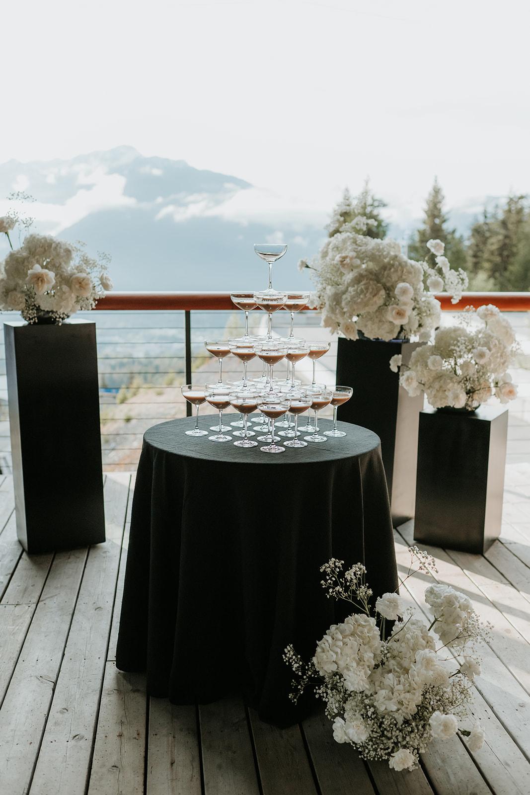 Champagne tower on a black table with white flowers, outdoor mountain view.