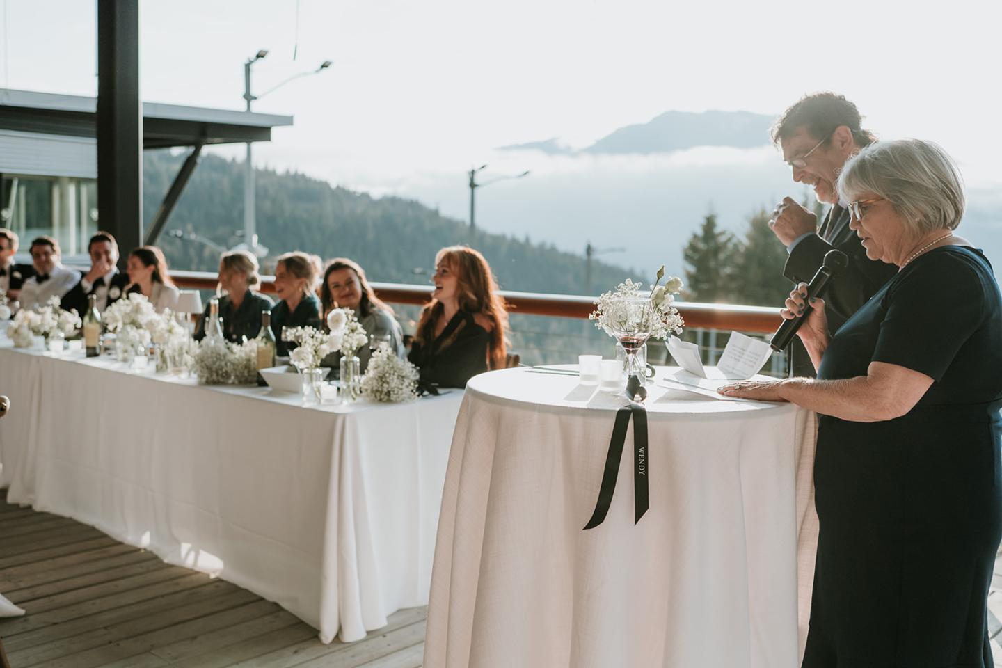 Wedding reception speech on a balcony with mountain views, guests seated at decorated tables.