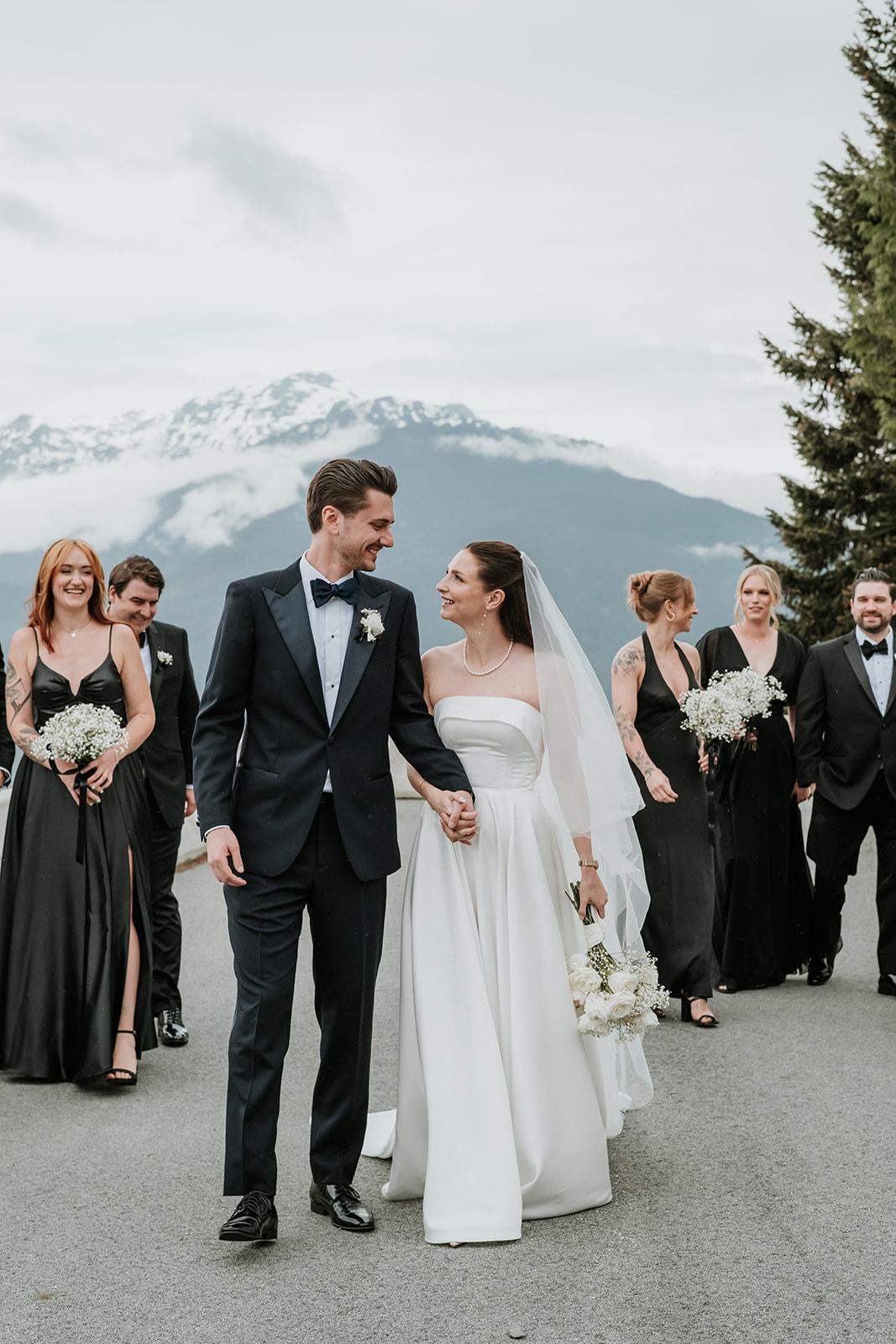 Bride and groom walking with wedding party, snow-capped mountains in background.