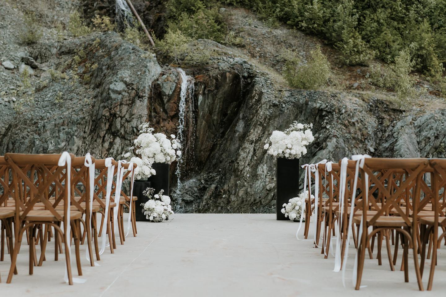Outdoor wedding setup with chairs facing a rocky waterfall, decorated with white flowers.