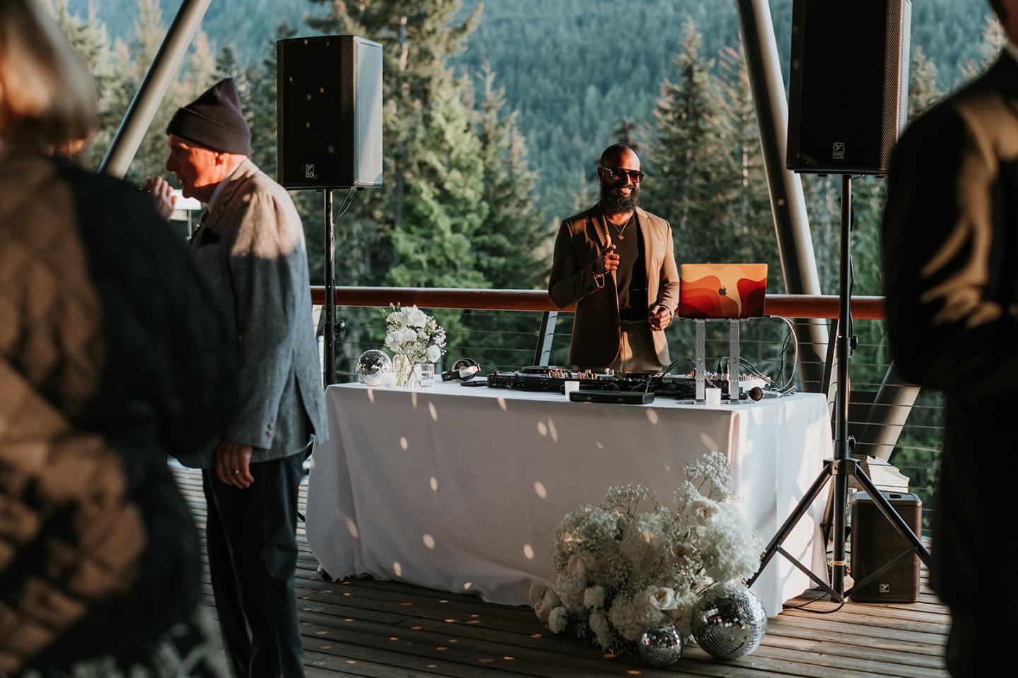 DJ stands behind a table at an outdoor event, with trees in the background.
