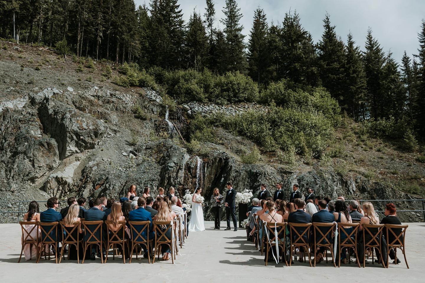 Outdoor wedding ceremony with guests seated, surrounded by rocky landscape and trees.