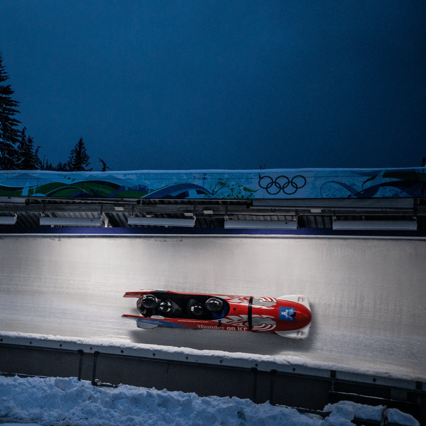 Red bobsled racing down an icy track at night with trees and Olympic rings in the background.