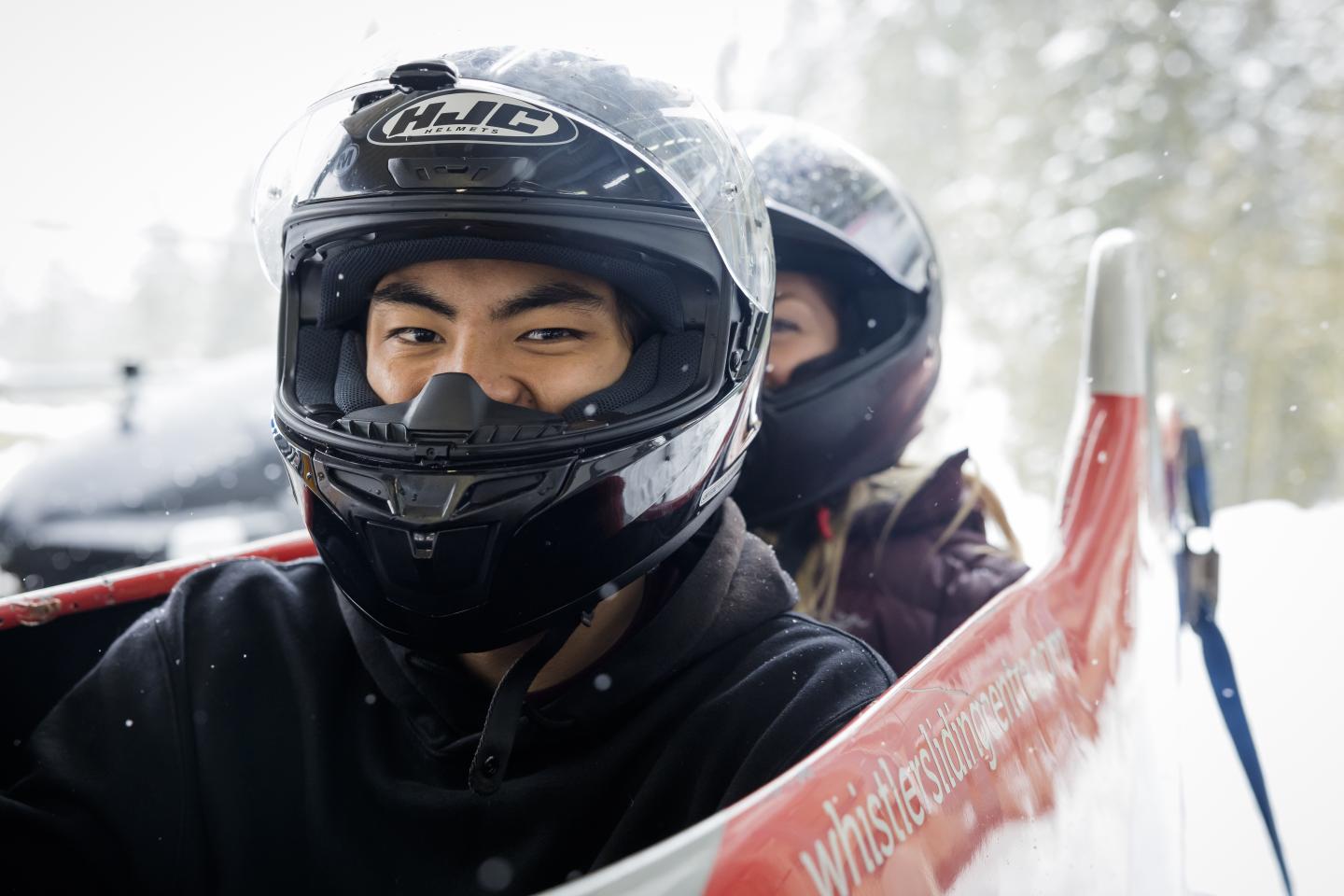 Two people wearing helmets in a bobsled on a snowy day.