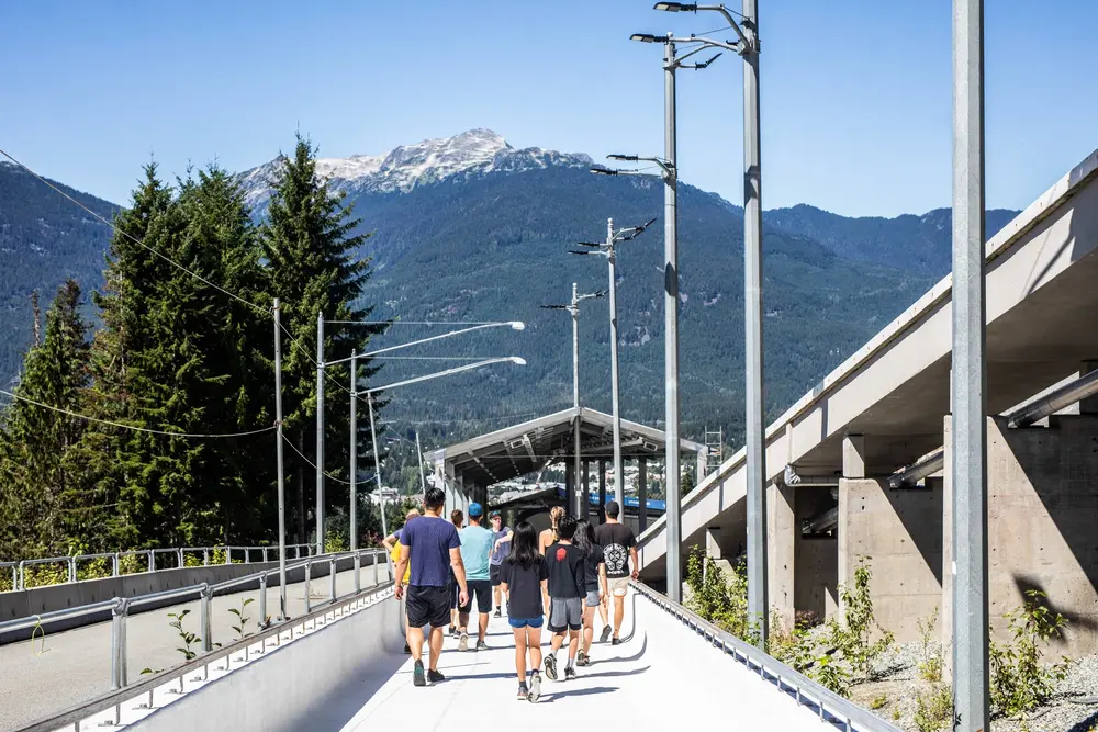 People walking on a sunny path with mountains and trees in the background.