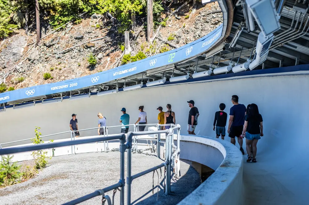 People walking on a bobsled track surrounded by trees in bright daylight.