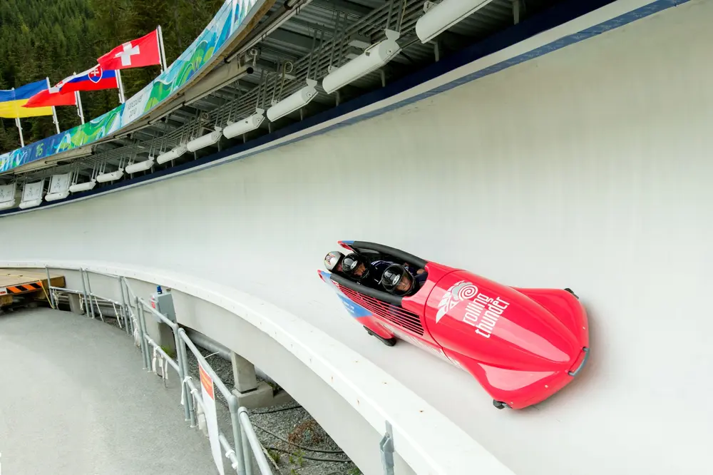 Red bobsled racing down an icy track, flags above.