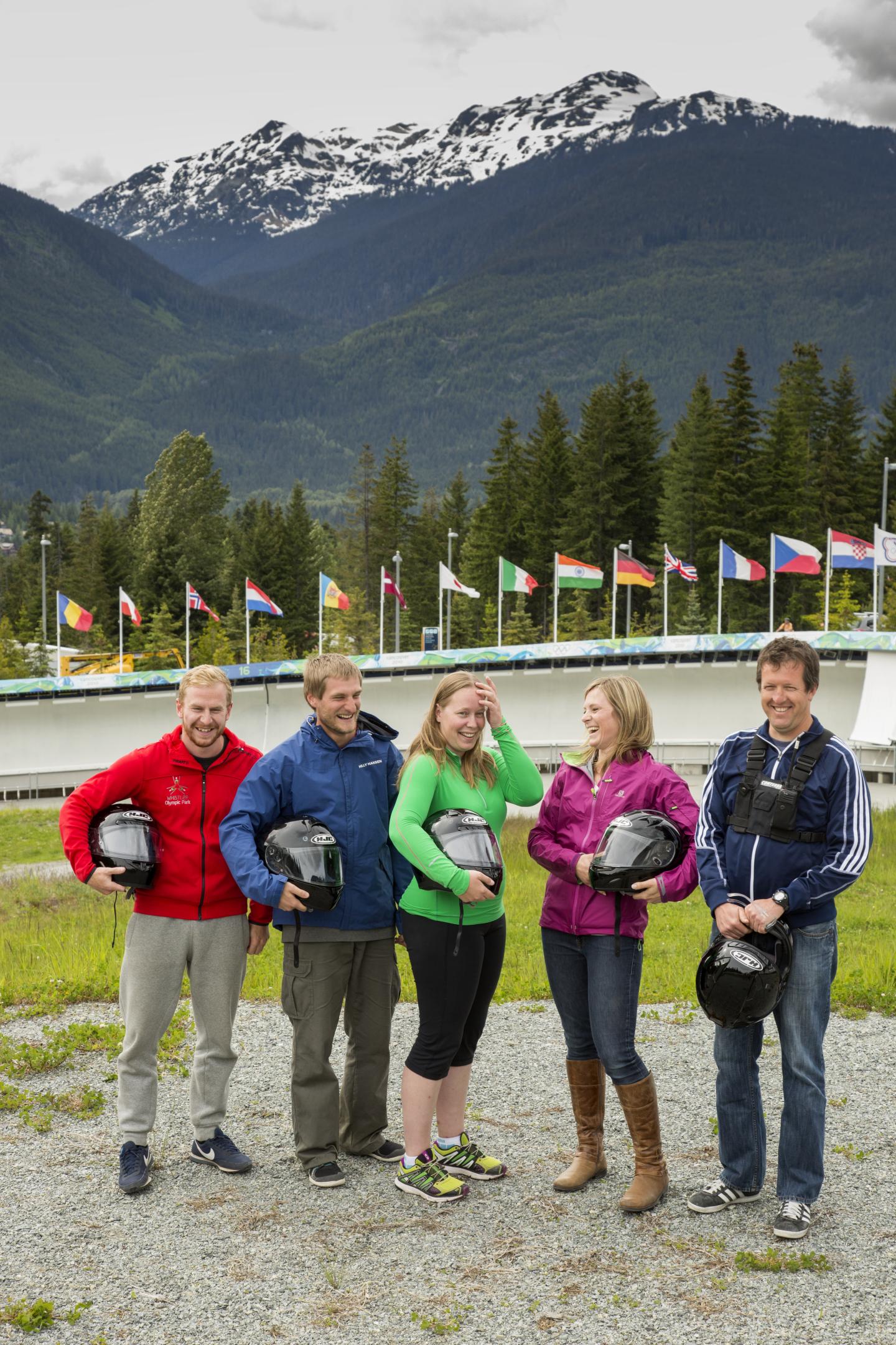 Five people with helmets smiling, standing before mountains and flags.