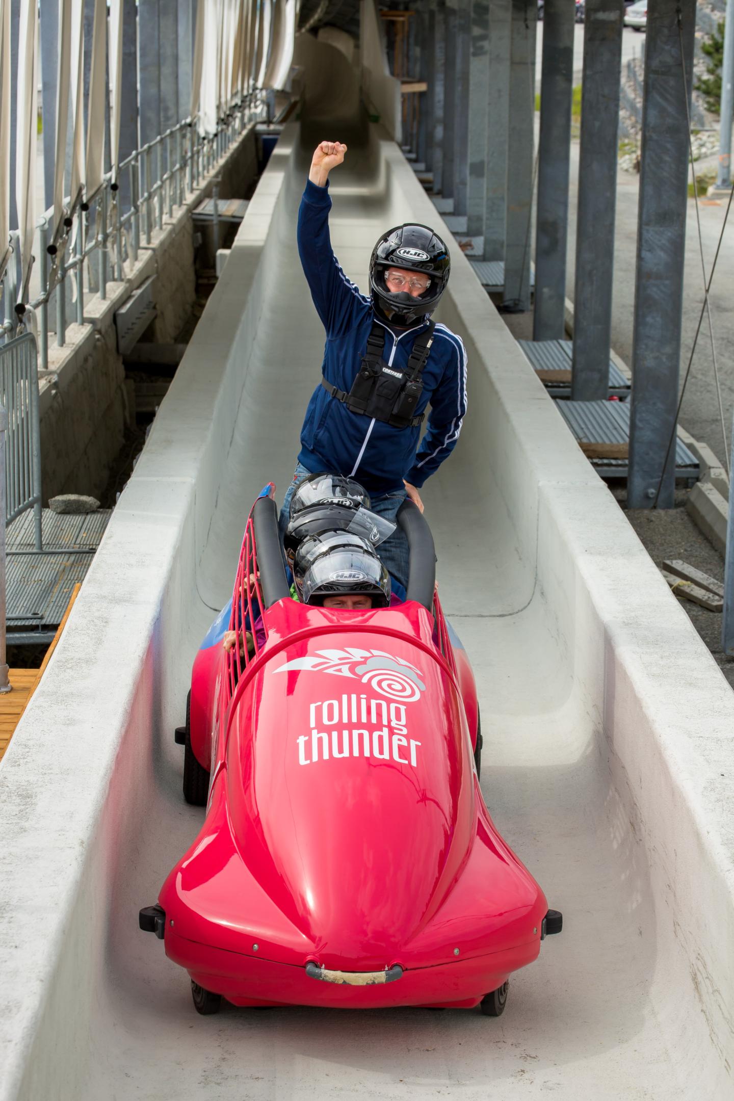 Person in helmet cheers while riding a red bobsled on a track.